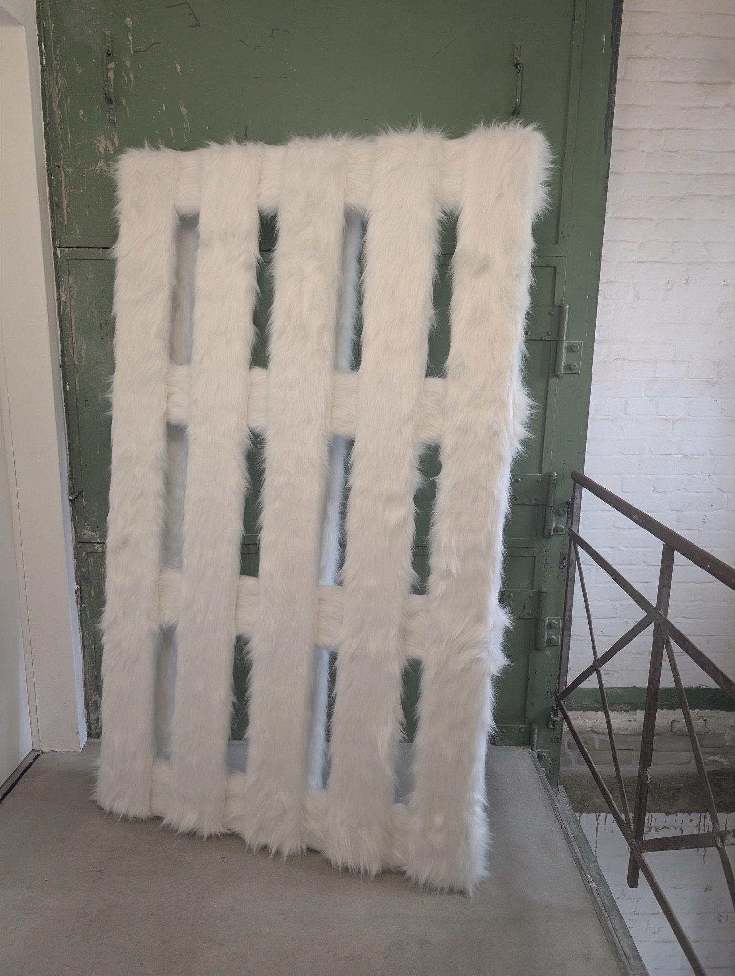 Mit weißem Kunstfell bezogene Palette an einer genieteten grünen Stahltür, Treppenpodest der Konrad Fischer Galerie